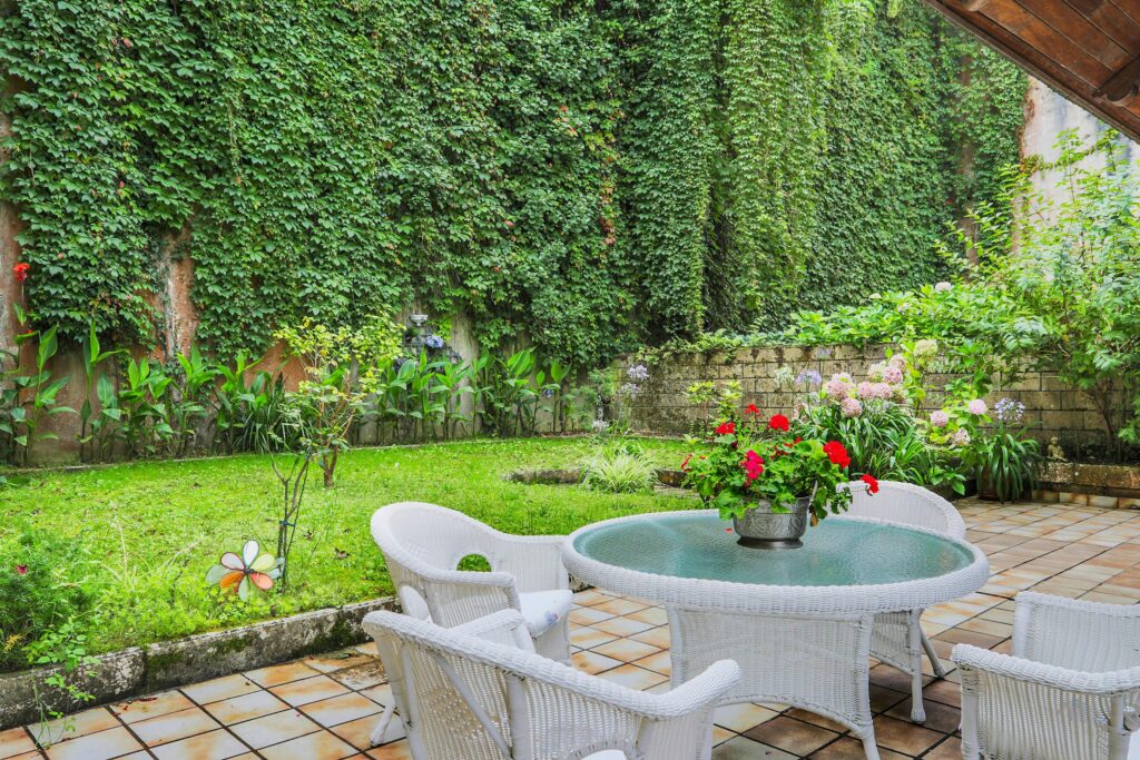 A charming garden patio with white wicker furniture surrounded by lush greenery and climbing vines.
